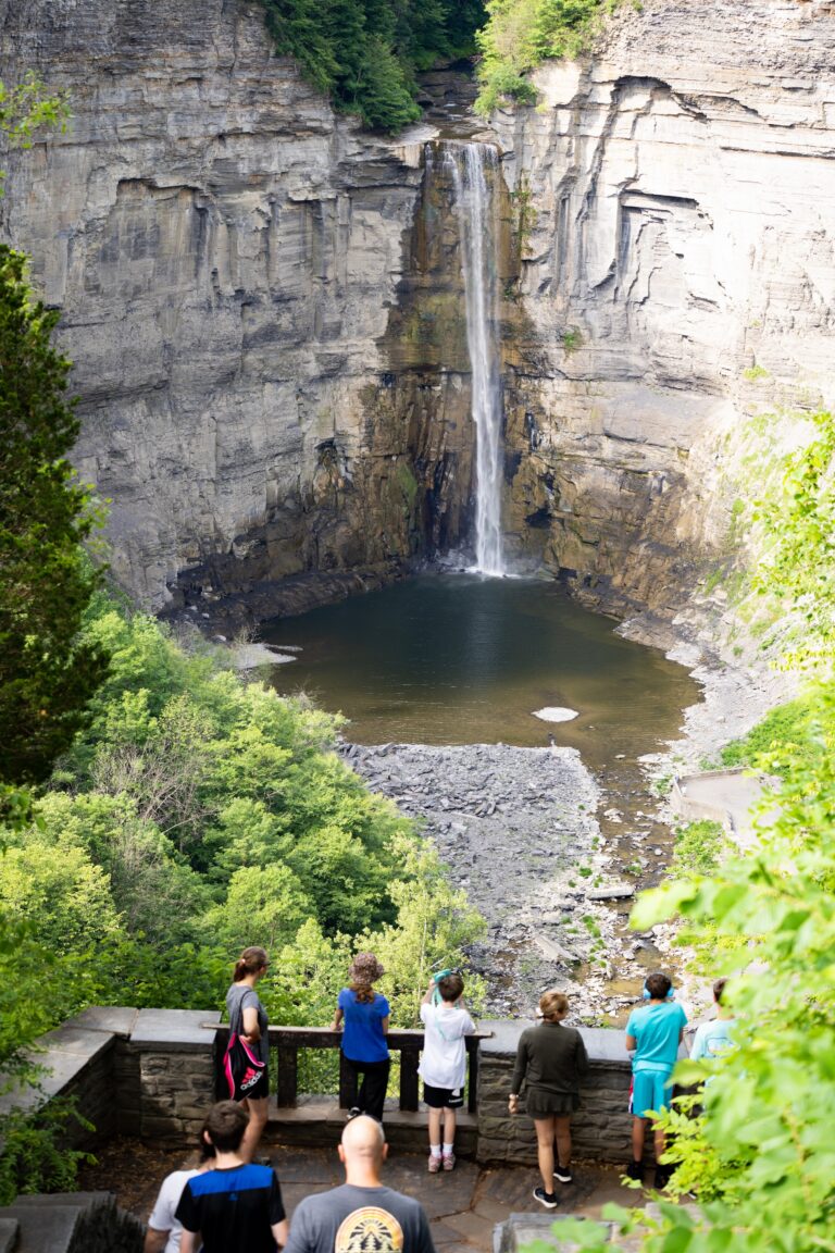 People stand at a stone overlook, watching a tall waterfall flow into a pool below, surrounded by steep cliffs and green trees.
