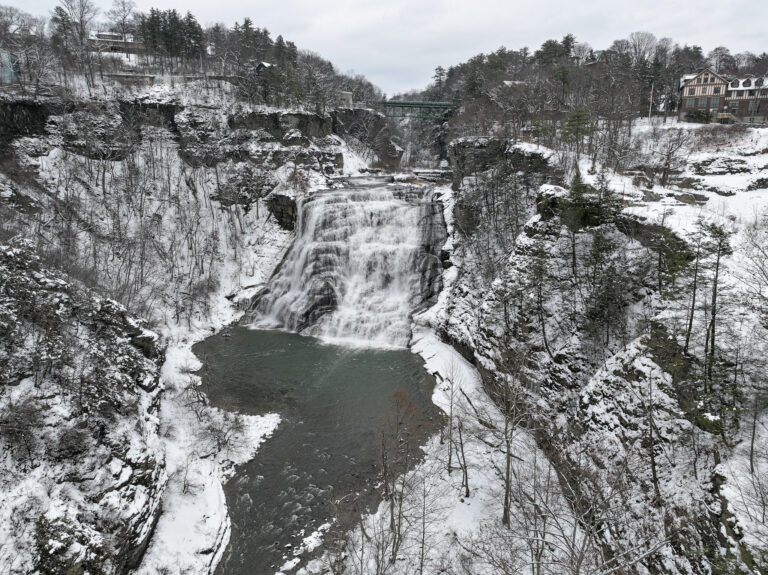 Wide waterfall flowing between snowy cliffs, with a green bridge above and snow-covered trees and buildings in winter.