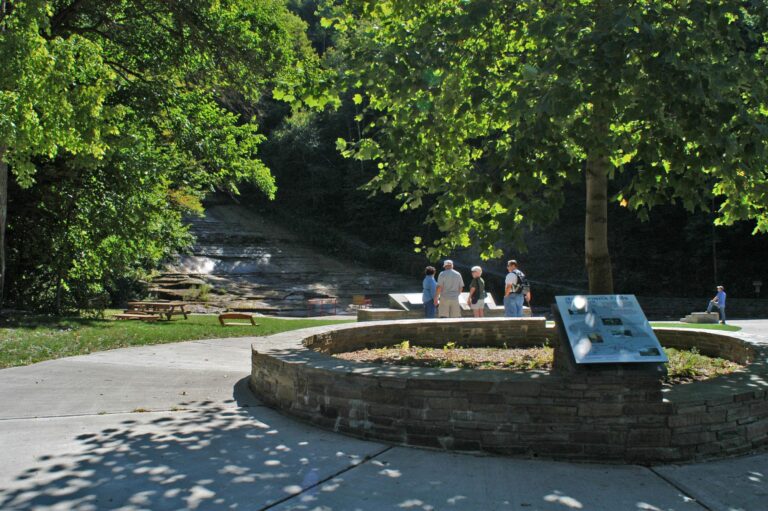 A group of people standing by an informational sign and stone planter in a sunny park with trees, picnic tables, and a small waterfall in the background.