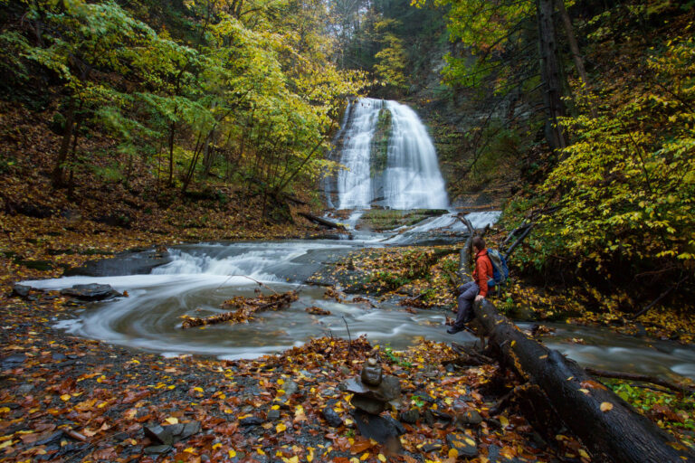 Person in red jacket sits on fallen tree, facing a waterfall with autumn trees; swirling water flows over rocks in the foreground.