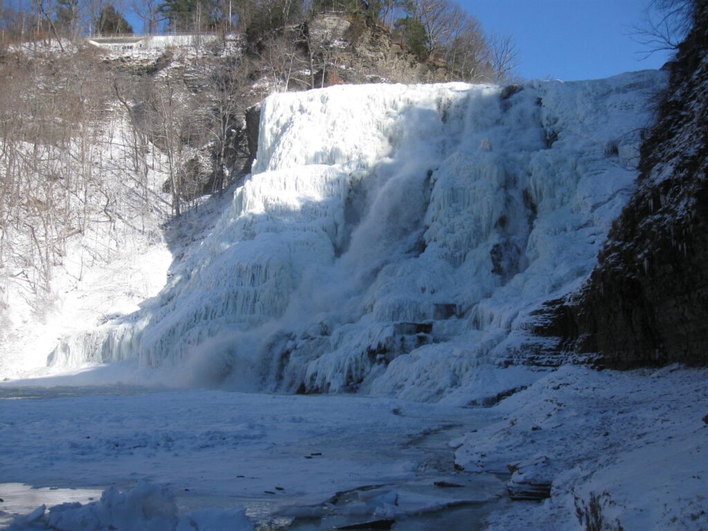 Frozen waterfall covered in thick ice, surrounded by snow and leafless trees under a clear blue sky.