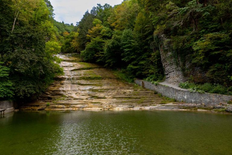 A calm pool at the base of a rocky slope, bordered by dense green trees and stone walls, beneath a partly cloudy sky.