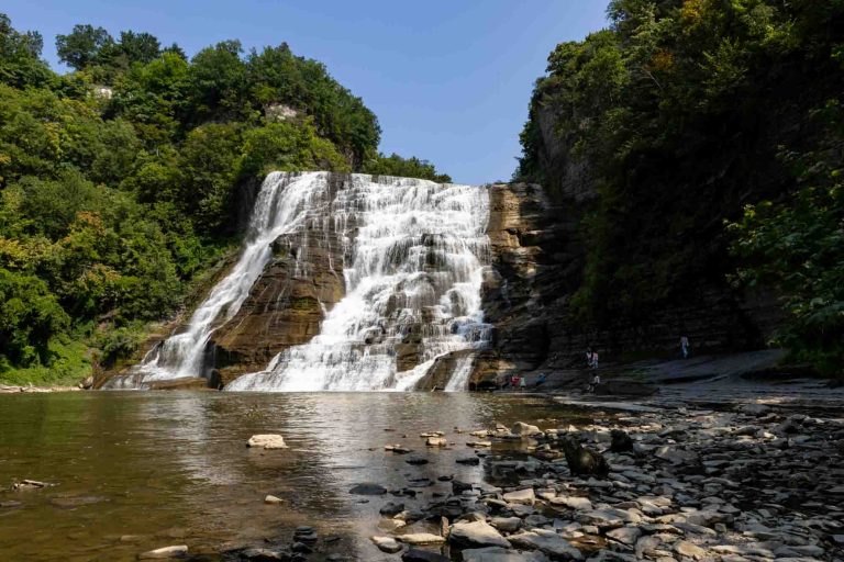 Wide waterfall flows over rocks into a shallow pool, surrounded by green trees and banks. Several people stand at the water's edge.