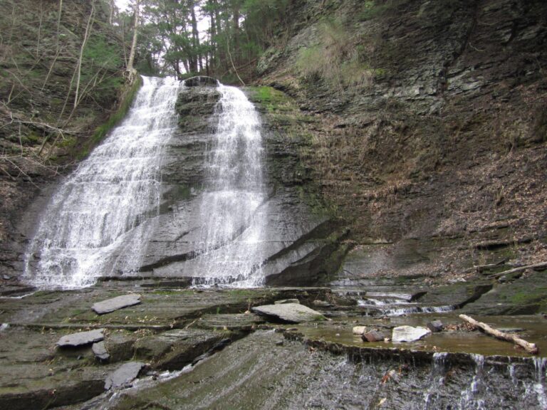 Waterfall flowing over layered rocks into a shallow pool, surrounded by moss-covered stones and trees.