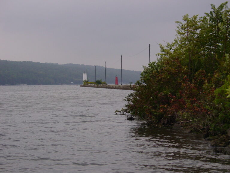 Cloudy lakeshore with a stone jetty extending into the water; two small lighthouses, one white and one red, stand at the jetty’s end. Green shrubs and trees line the shore in front.