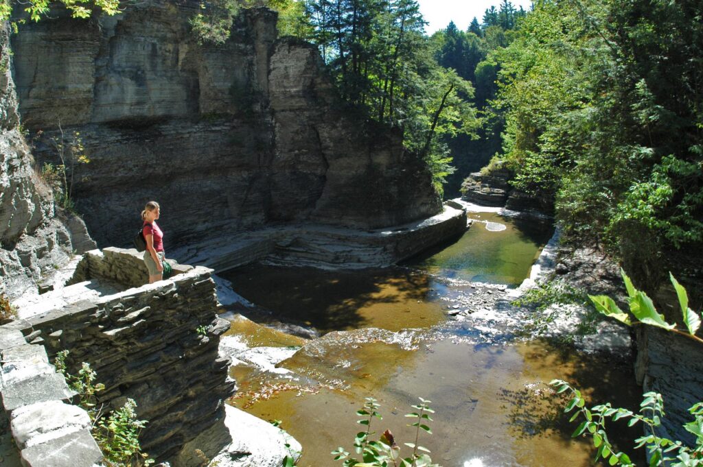 Person standing on a stone ledge above a rocky gorge with shallow water, surrounded by tall cliffs and green trees in bright sunlight.