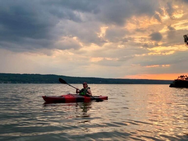 Person kayaking on a calm lake at sunset, with orange-pink sky and clouds reflected in the water; tree-covered hills in the background.