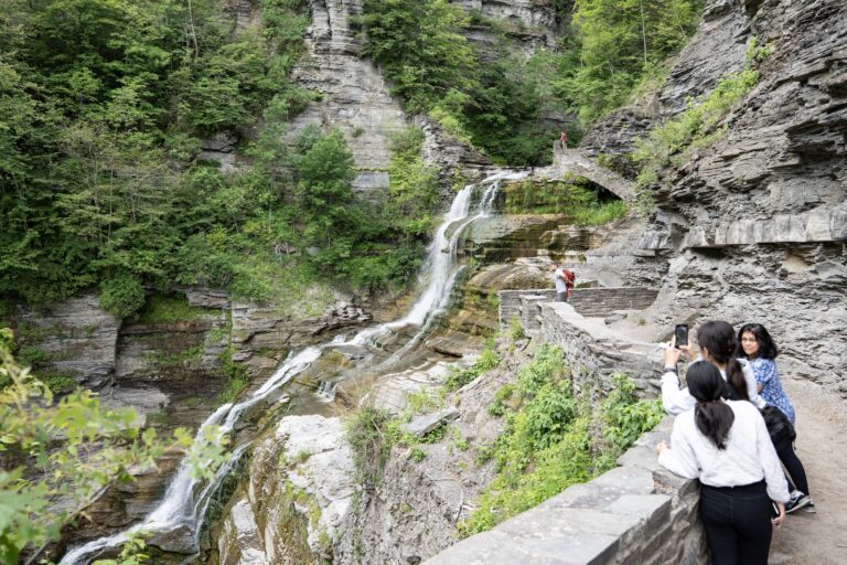 People stand on a stone path beside a waterfall and green cliffs in a gorge, with rocky walls and dense trees.