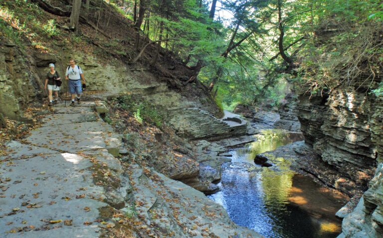 Two hikers walk on a rocky trail next to a narrow gorge with layered cliffs, a flowing stream, and dense green trees.