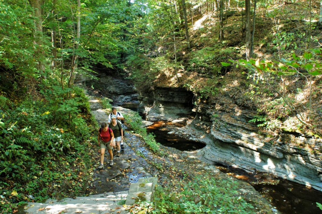 Three people hiking on a rocky trail through a green forest next to a narrow gorge with layered rock walls and small pools, under dappled sunlight.