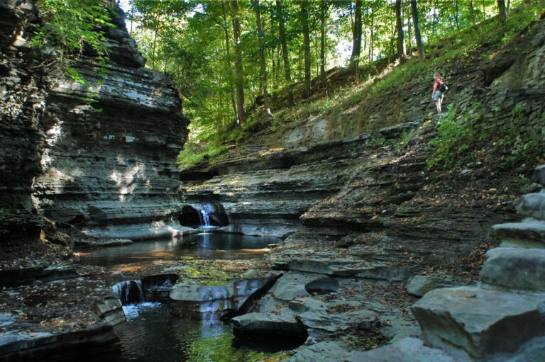 Hiker on a rocky trail above stone cliffs and a stream with waterfalls, surrounded by green forest.
