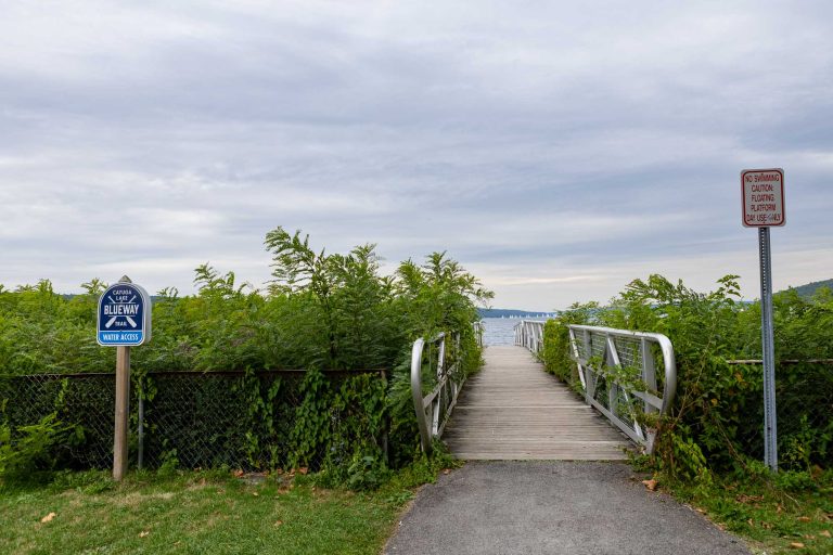 Wooden boardwalk through green bushes to water under cloudy sky; signs show blueway access, no swimming, diving, or fishing.