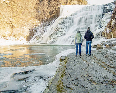Two people in winter jackets stand on a rocky, icy shore, facing a partially frozen waterfall with sunlight shining on the scene.