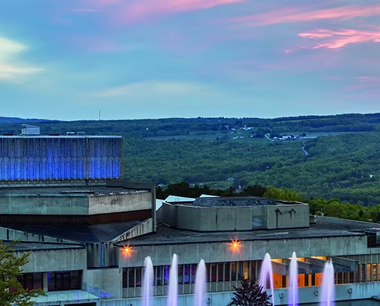 Modern concrete building with blue accent lights, green hills and a colorful sunset sky behind, and water fountains in the foreground.