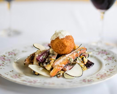 Gourmet dish with a crispy fried ball atop roasted vegetables, garnished with sauce and thinly sliced radishes on a floral plate; wine glasses are in the background.