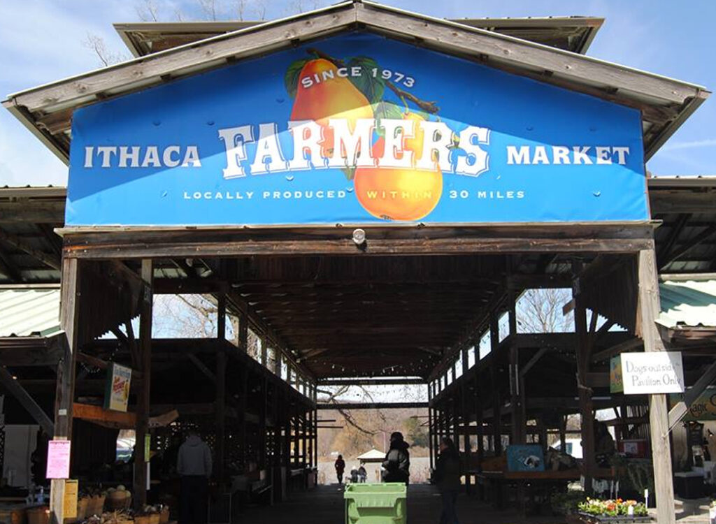 Blue Ithaca Farmers Market banner hangs above an open-air market entrance, with people and vendor stalls visible inside. The sign says Since 1973 and Locally produced within 30 miles.